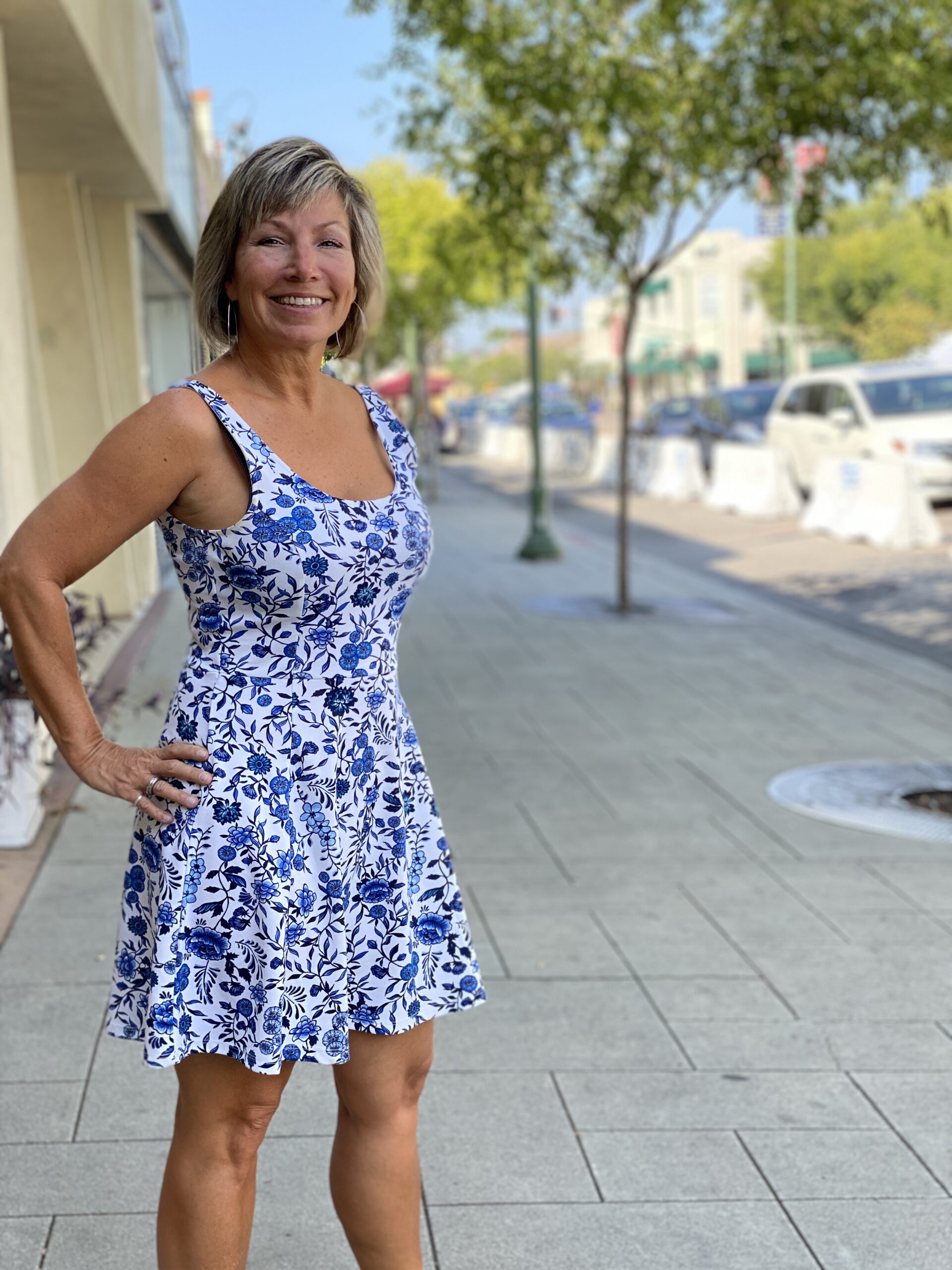 A woman wearing a blue and white dress stands gracefully on a sidewalk, surrounded by a vibrant urban setting.